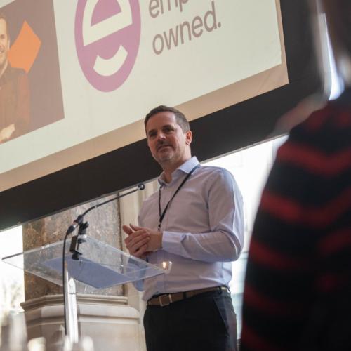 James de la Vingne from the Employee Ownership Association speaking at the Aspire Banking Hall in front of a projector screen.