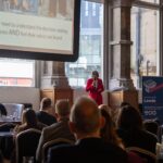 Noo Jones from Athena Professional speaking at the Aspire Banking Hall in front of a projector screen.