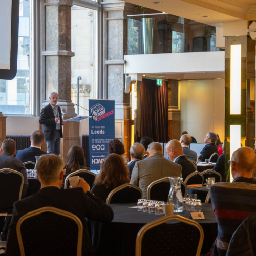 Robert Camp, conference chair, speaking at the Aspire Banking Hall in front of a projector screen.